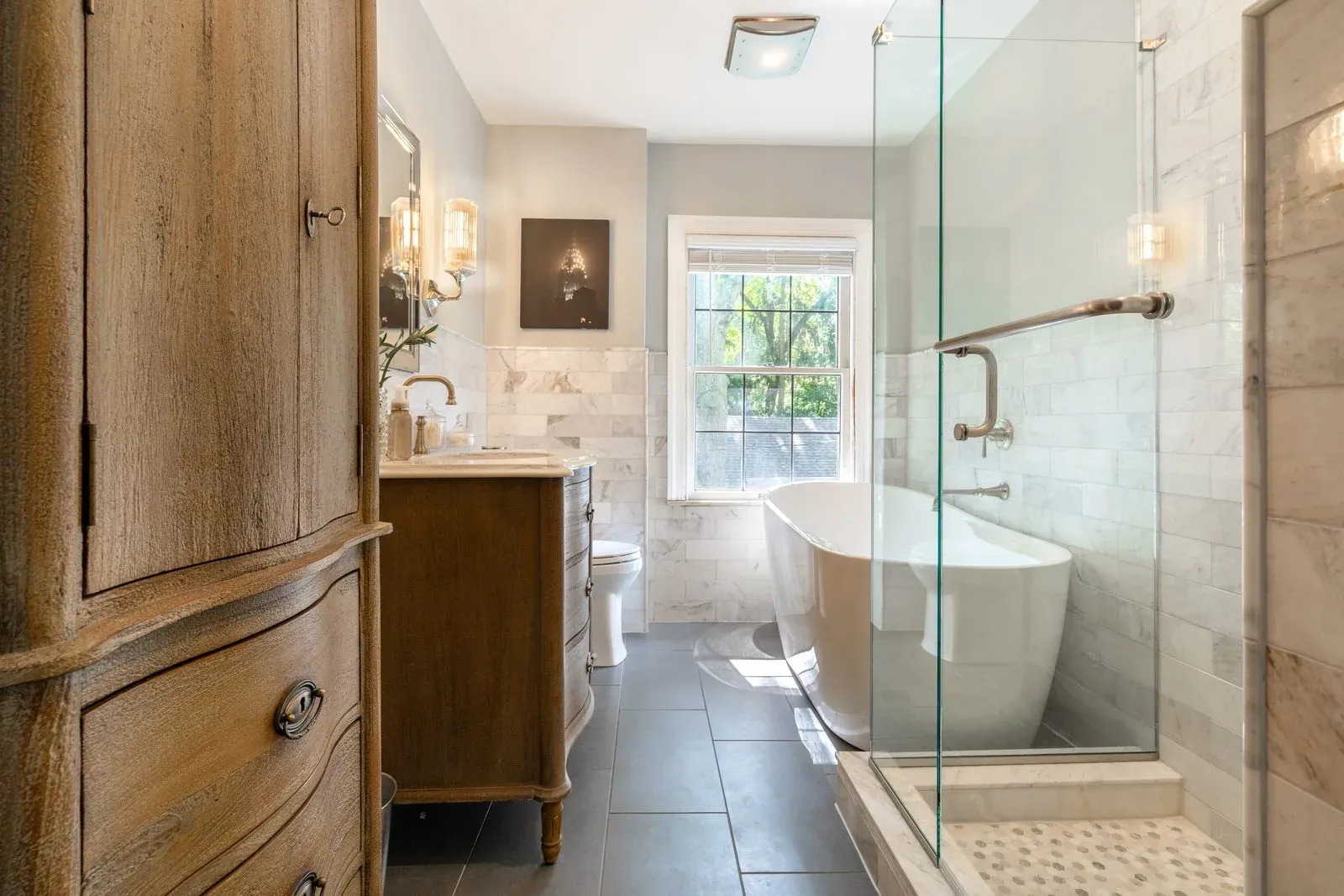 Full bathroom remodel with a curved stained wood vanity, marble subway tile, a freestanding tub under the window and a frameless glass shower