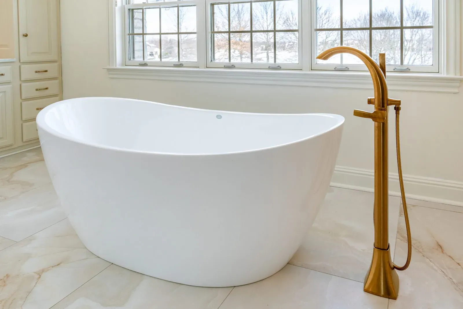Replacement freestanding soaking tub with a brushed gold floor-mounted filler, set under a bank of windows on marble-look tile