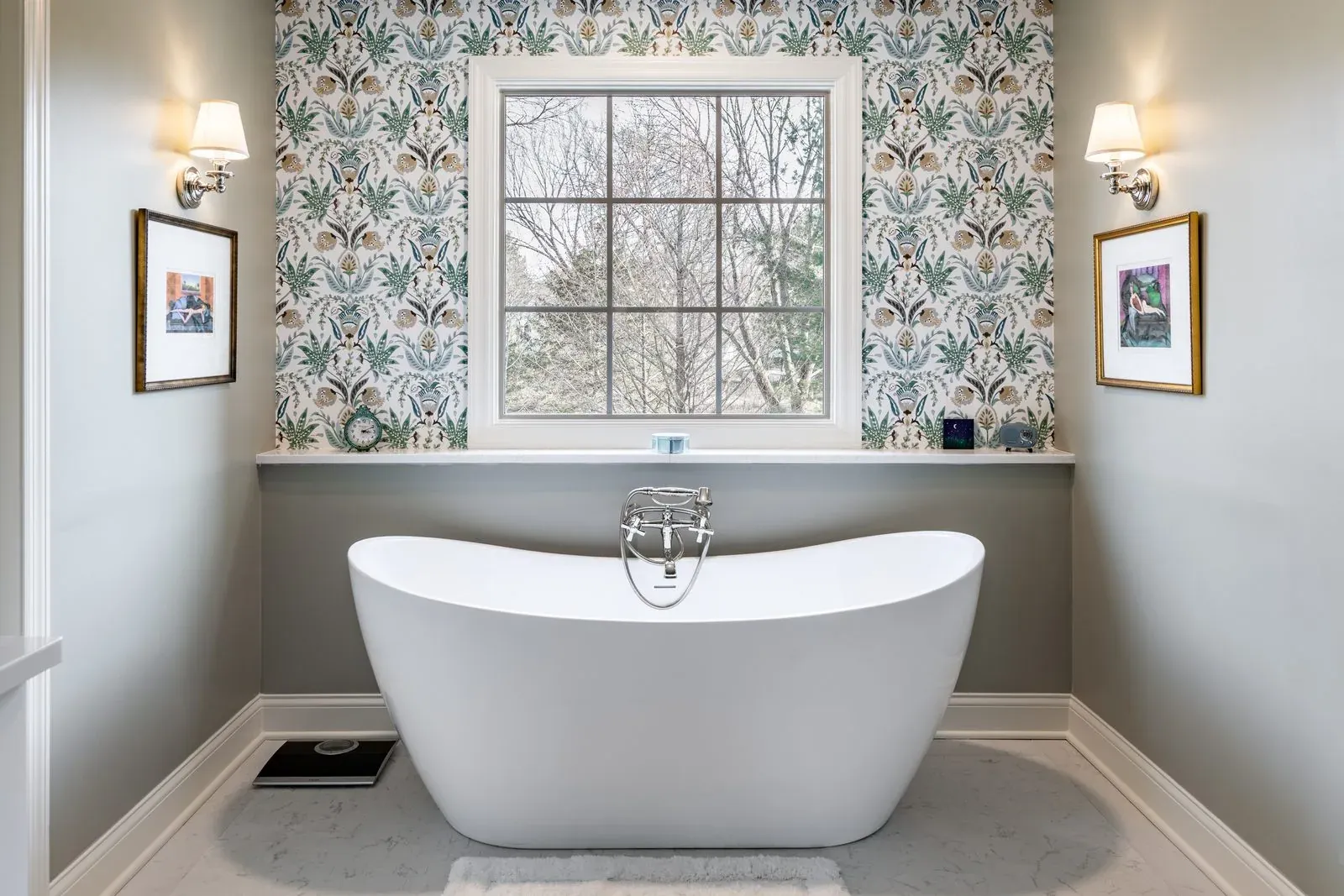 Freestanding slipper tub beneath a window on a botanical wallpaper accent wall, flanked by polished nickel sconces and framed art