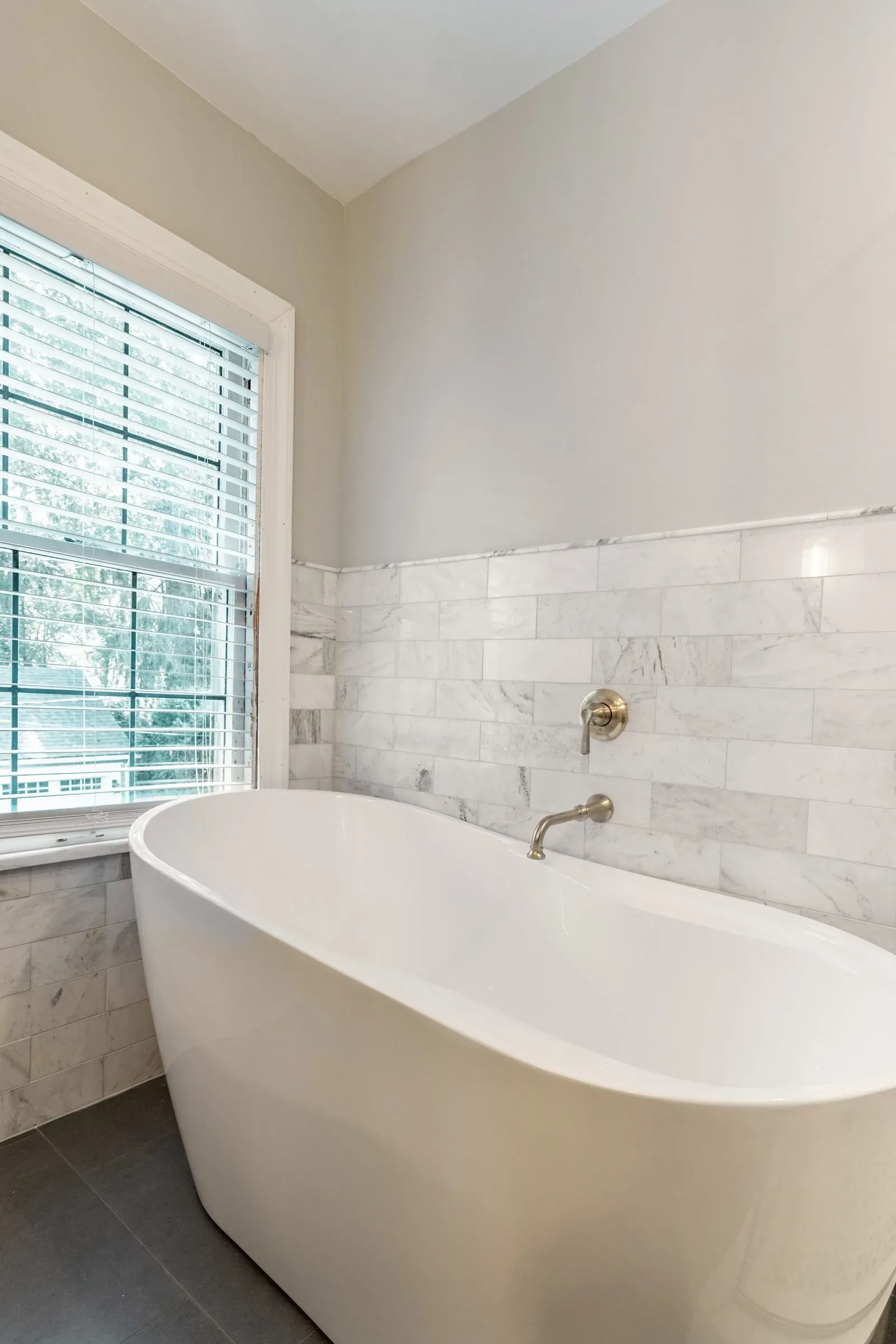 Freestanding tub against a marble subway tile wall with a wall-mounted brushed gold tub filler and a shuttered window alongside