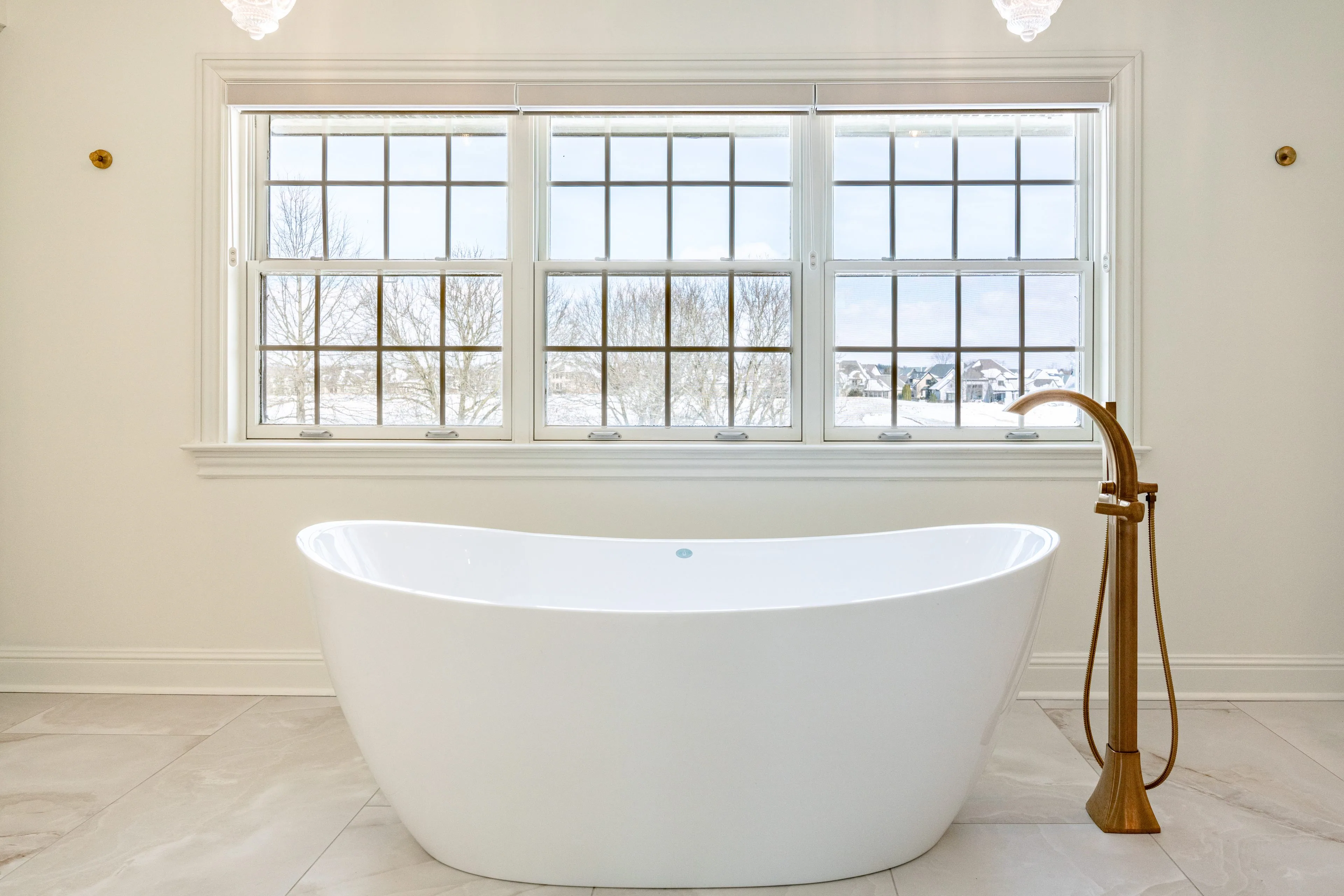 White freestanding soaking tub centered under a bank of windows with a brushed gold floor-mounted tub filler and marble-look tile flooring