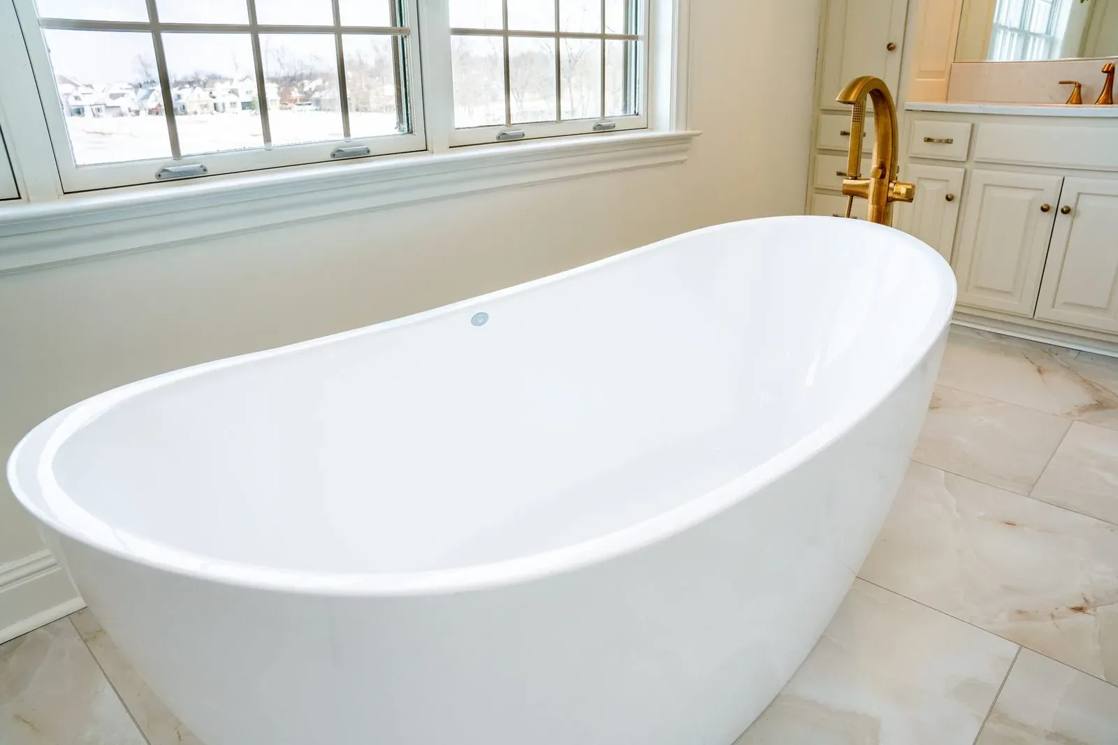 Replacement freestanding soaking tub under a window bank, with a brushed gold floor-mounted filler on marble-look tile flooring