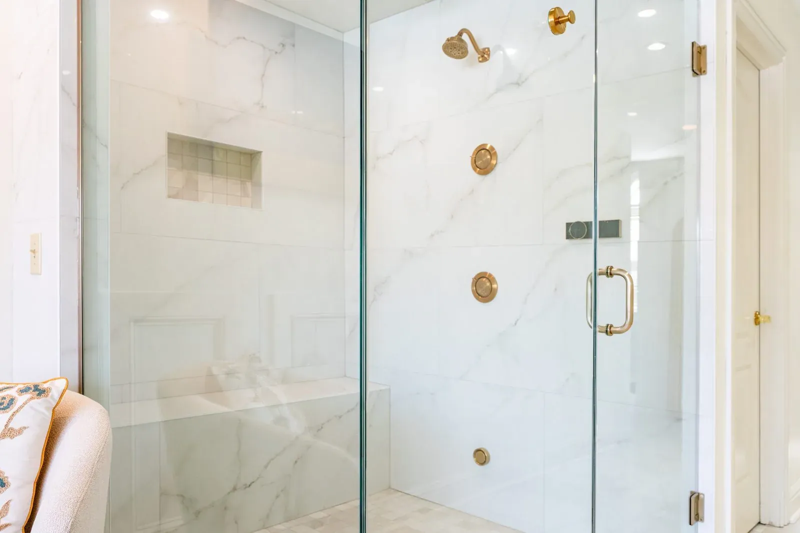 Frameless glass shower tiled floor to ceiling in marble-look porcelain, with brushed gold body sprays, a recessed niche and a built-in bench