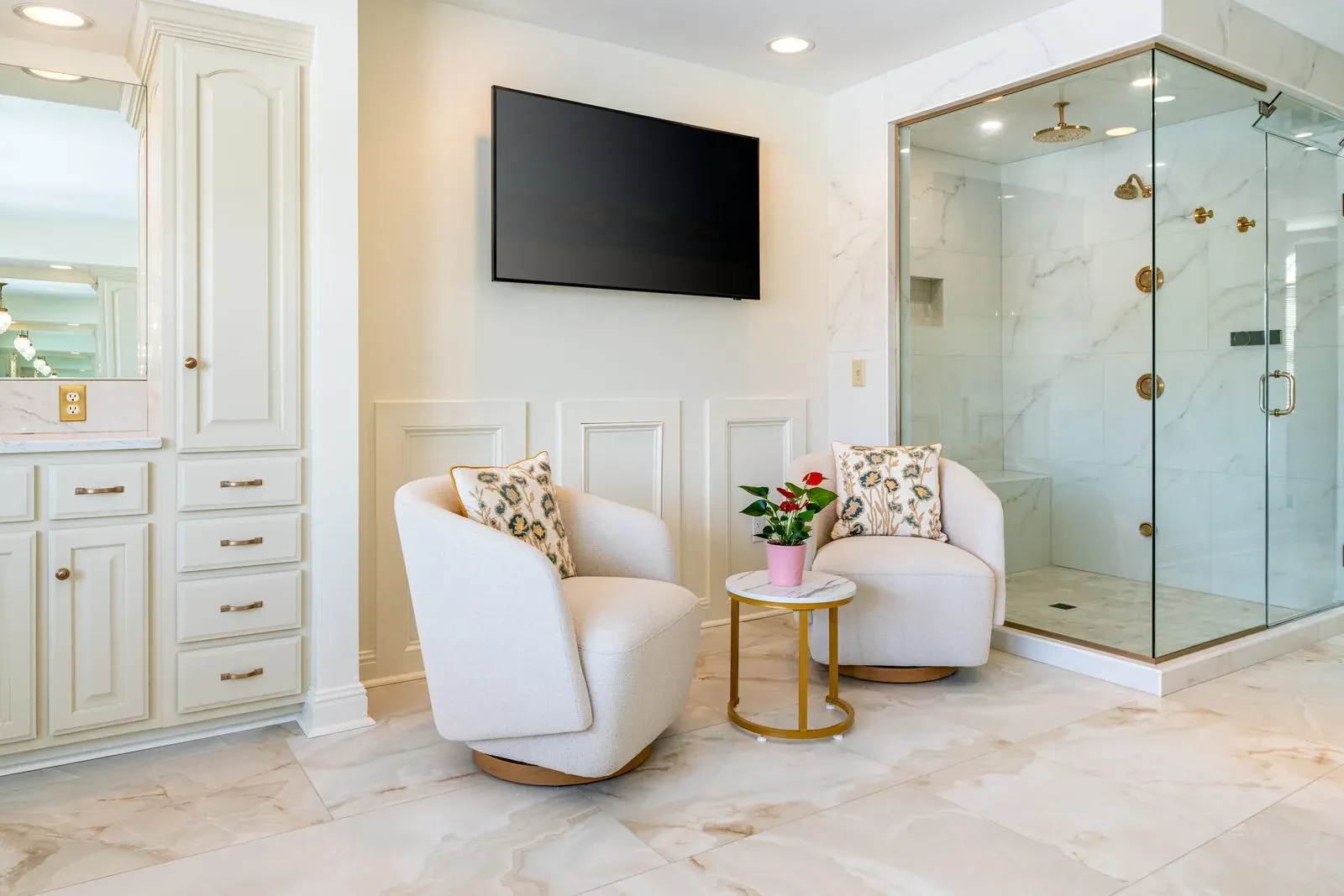 Primary bathroom remodel with a glass marble-tiled shower, cream inset cabinetry, a wall-mounted television and two upholstered swivel chairs