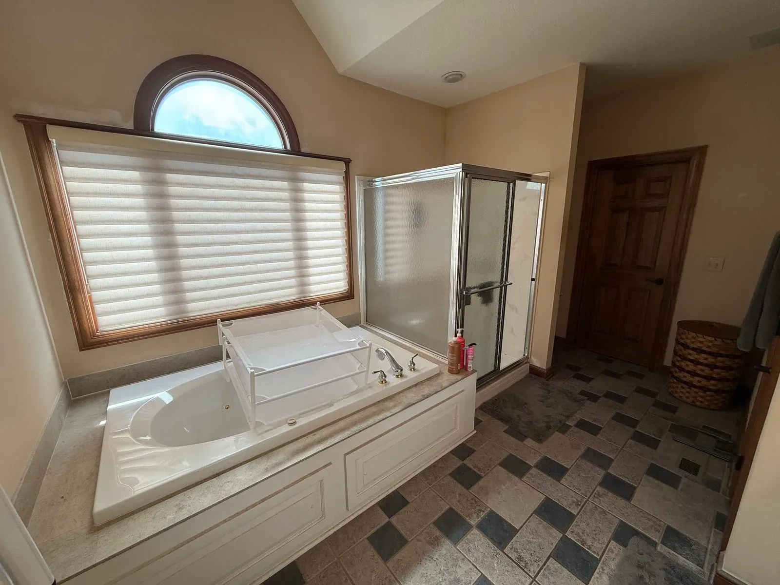 Bathroom before the conversion, with an oversized jetted tub set into a tiled deck below an arched window, a framed glass shower enclosure and dark checkerboard tile flooring