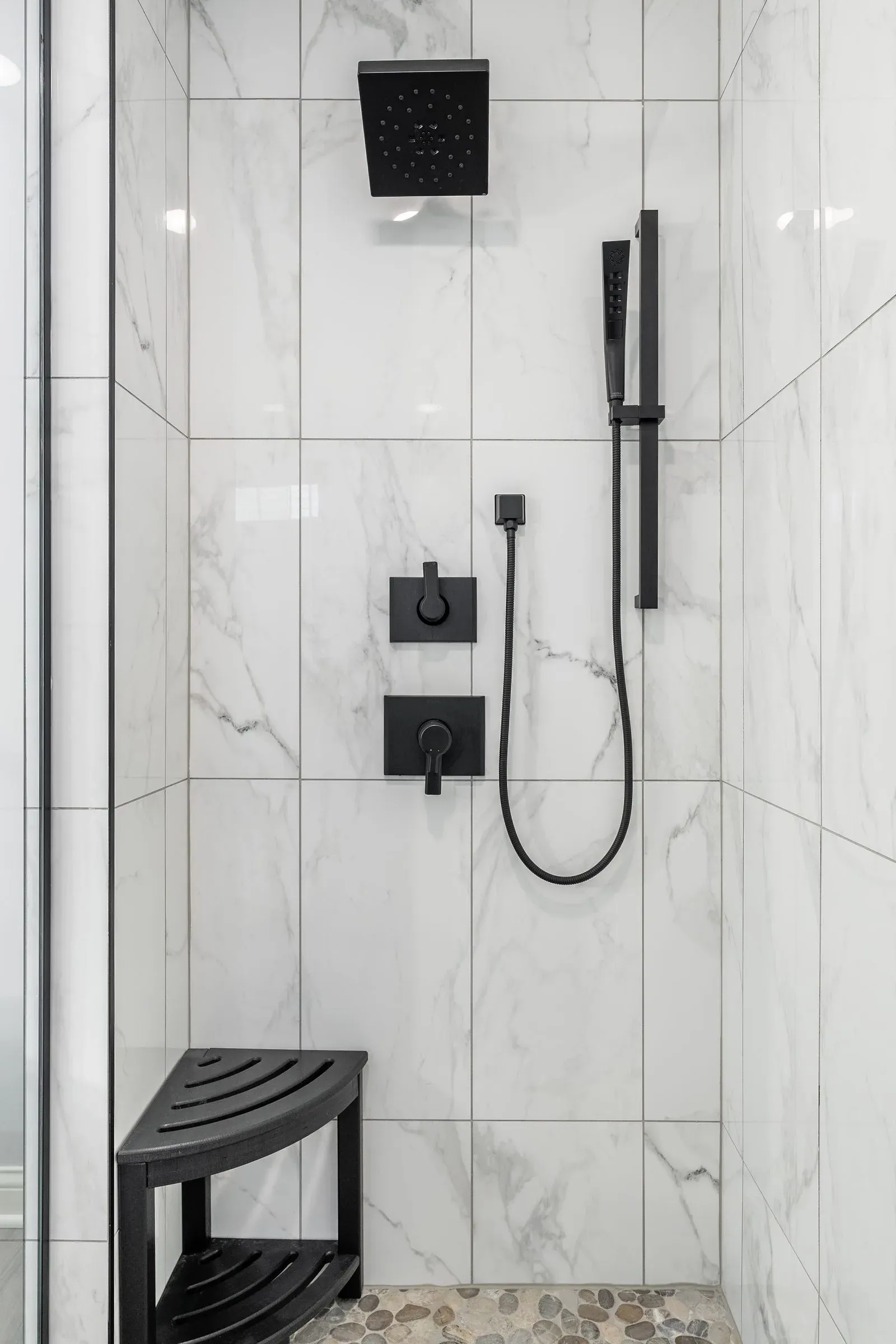 Walk-in shower tiled in marble-look porcelain with matte black controls, a hand shower on a slide bar and a black corner seat