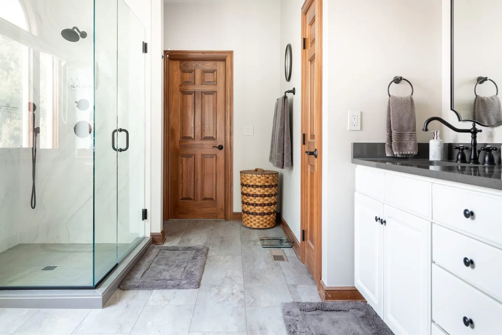Newly installed frameless glass shower with marble-look wall tile and matte black fixtures, beside a white double vanity