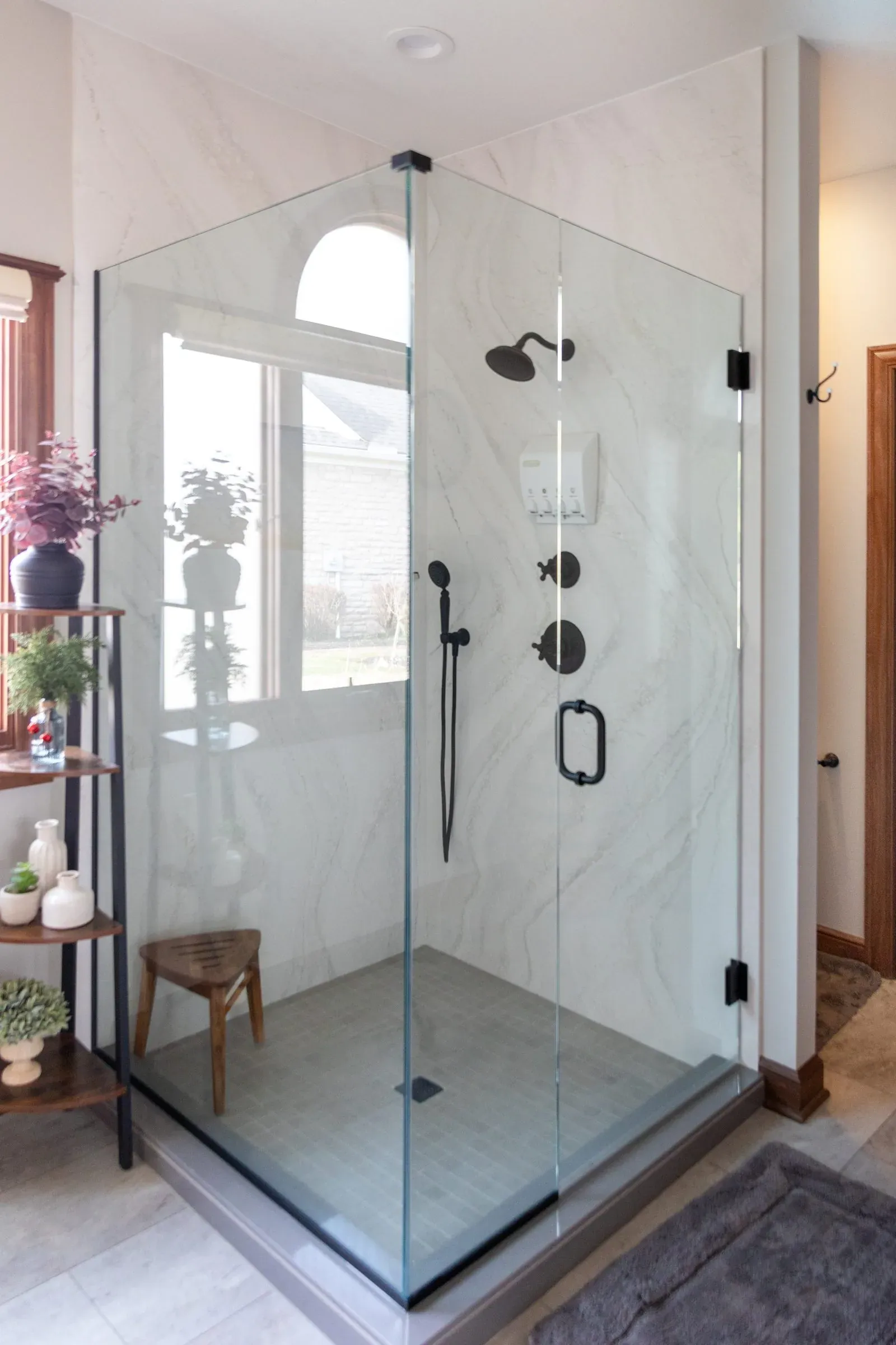 Corner glass shower tiled in marble-look porcelain with a matte black rain head, hand shower and body sprays, beside a window